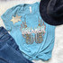 Dreamer Screen Print Heat Transfer, colorful graphic t-shirt with feather design, featuring the word DREAMER, laid on white faux fur with denim jeans and a black hat.