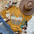 Dreamer Screen Print Heat Transfer, colorful graphic of wings on a mustard yellow T-shirt with boho accessories, hat, jewelry, and cowhide pattern to showcase the product design and style.