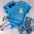 Road Trippin POCKET Screen Print Heat Transfer, blue t-shirt with mountain and van graphic, paired with distressed denim shorts, white sneakers, sunglasses, and a small flower arrangement.