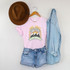 Got That Sunshine In My Pocket Screen Print Heat Transfer T-Shirt, pink casual t-shirt with mountain and sunrise graphic design, paired with denim shorts and a light blue denim shirt, brown wide-brim hat hanging on a hanger on a white background.