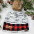 Not Made For Winter TAN Leather Patch, knitted winter beanie hat with a faux fur pom-pom, white, black, and red color stripes, on a white surface with pine branches and pinecones for winter decoration.
