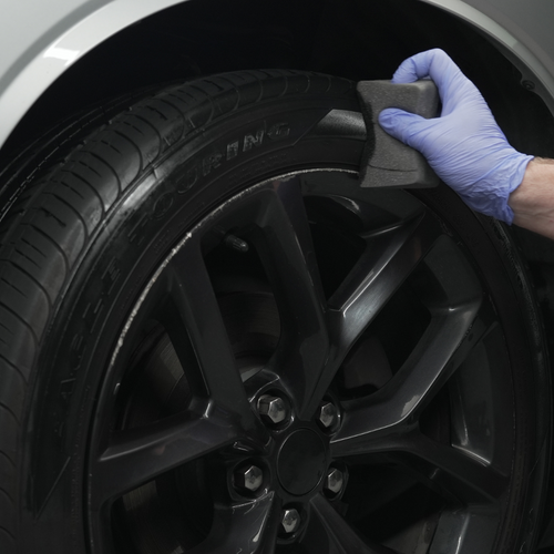 Gloved hand applying Wipe New Midnight Black Tire Shine to black car tire with foam applicator