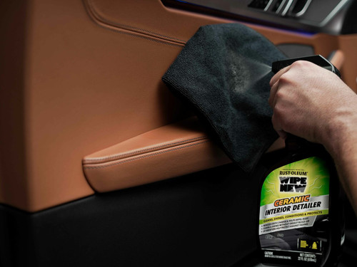 The scene takes place near the interior of a car door, showing Wipe New Ceramic Interior Detailer being sprayed onto a plush microfiber cloth. A fine mist of cleaner is dispensed from the nozzle, preparing the cloth to tackle various interior surfaces. The bottle's eye-catching label and prominent branding stand out, showcasing its ability to clean, shine, and condition. The car's pristine interior in the background underscores the product's effectiveness in revitalizing and protecting materials like leather, plastic, vinyl, and rubber.