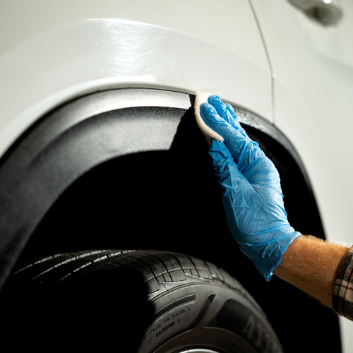 A person wearing a blue glove cleans a car's wheel arch with a sponge.