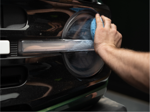 A person polishing a car's front grille with a blue cloth, showing reflection on the surface.