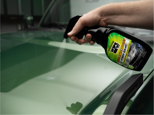A hand holding a bottle of ceramic boost spray, applying it to a shiny green car hood.