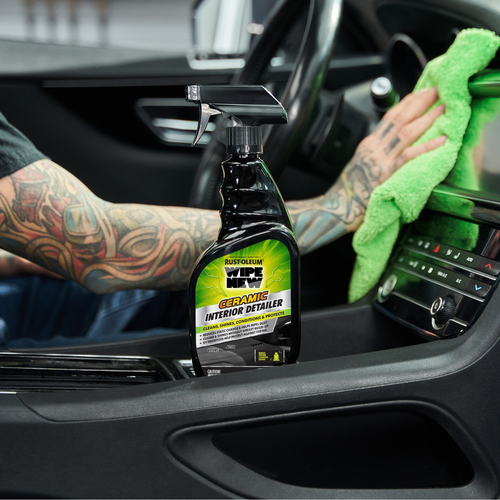 A person with tattooed arms wipes car interior with a green cloth. A spray bottle of Rust-Oleum Ceramic Interior Detailer is nearby.
