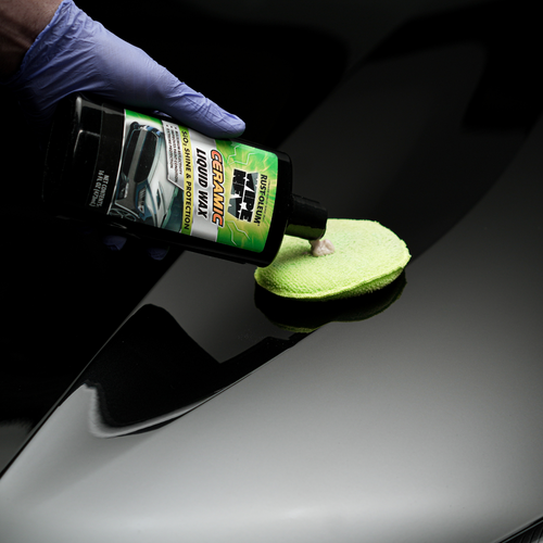 A gloved hand pouring ceramic liquid car wax onto a green microfiber cloth, with a shiny black car surface in the background.