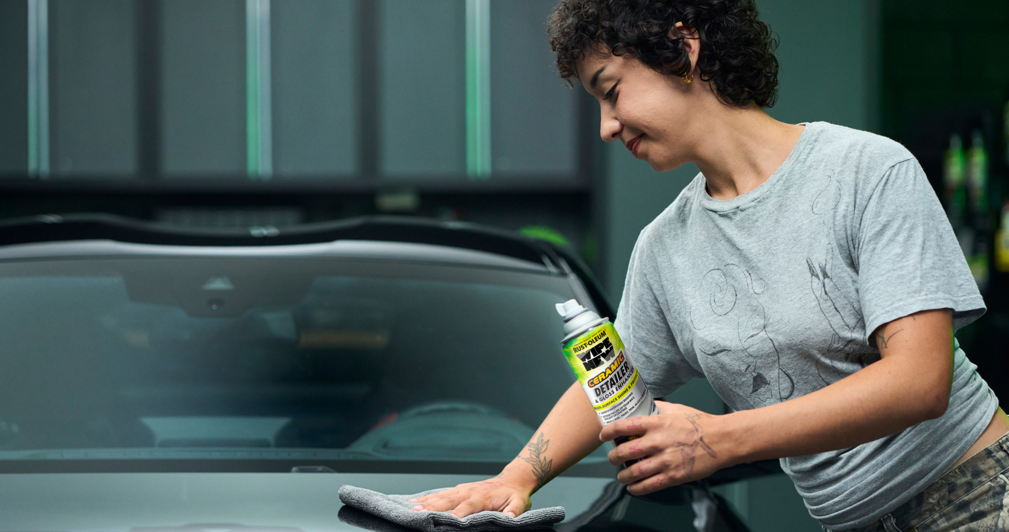 A young woman with short, curly dark hair is detailing the hood of a dark-colored car in an indoor garage setting. She holds a white and green Rust-Oleum Wipe New Ceramic Detailer & Gloss Enhancer spray can in her right hand, aimed at the car's hood, while her left hand presses a grey microfiber cloth against the surface. The car's hood is glossy and reflective, showcasing the product's effectiveness. The background is blurred, featuring dark walls with vertical green light strips and shelves with bottles, reinforcing the car care context. The woman wears a light grey t-shirt with subtle black patterns and camouflage pants, adding a casual yet professional vibe to the scene.