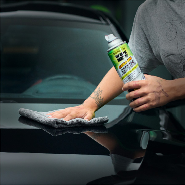 someone is detailing the hood of a dark-colored car in an indoor garage setting. She holds a white and green Rust-Oleum Wipe New Ceramic Detailer & Gloss Enhancer spray can in her right hand, aimed at the car's hood, while her left hand presses a grey microfiber cloth against the surface. The car's hood is glossy and reflective, showcasing the product's effectiveness. The background is blurred, featuring dark walls with vertical green light strips and shelves with bottles, reinforcing the car care context. The woman wears a light grey t-shirt with subtle black patterns and camouflage pants, adding a casual yet professional vibe to the scene.