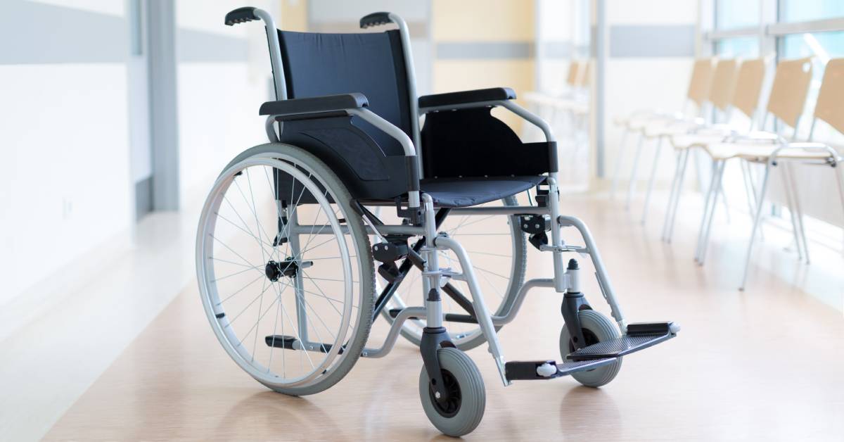 An empty wheelchair in a clear hospital hallway. The wheelchair has small caster wheels in front of the larger wheels.