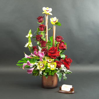 Together Forever is a  tall, sculptural and premium luxury arrangement featuring  red roses, yellow orchids, pink calla lilies, tropical greens, tall bamboo accents with vertical accents arranged in a modern container by Lexis Florist Houston.