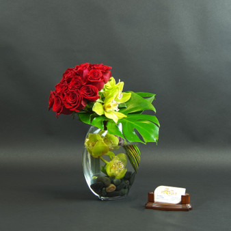 Love Struck is a very modern, sculptural, luxury rose  arrangement in a contemporary floatin and submerged style that can be ordered in 3 different sizes (small, medium and large). A large cluster of red roses featuring white cymbidium orchids and tropical green monstrea leaf arranged in a large clear glass bowl with river stones.