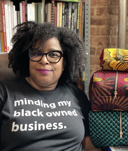 photo of black woman with an afro wearing a t-shirt that reads: Minding my own Black Business. Woman is seated next to three large and colorful zipper pouches.