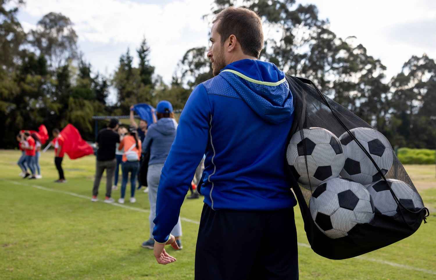 Photograph of a person in a blue jacket carrying a mesh bag filled with soccer balls on a grassy field.