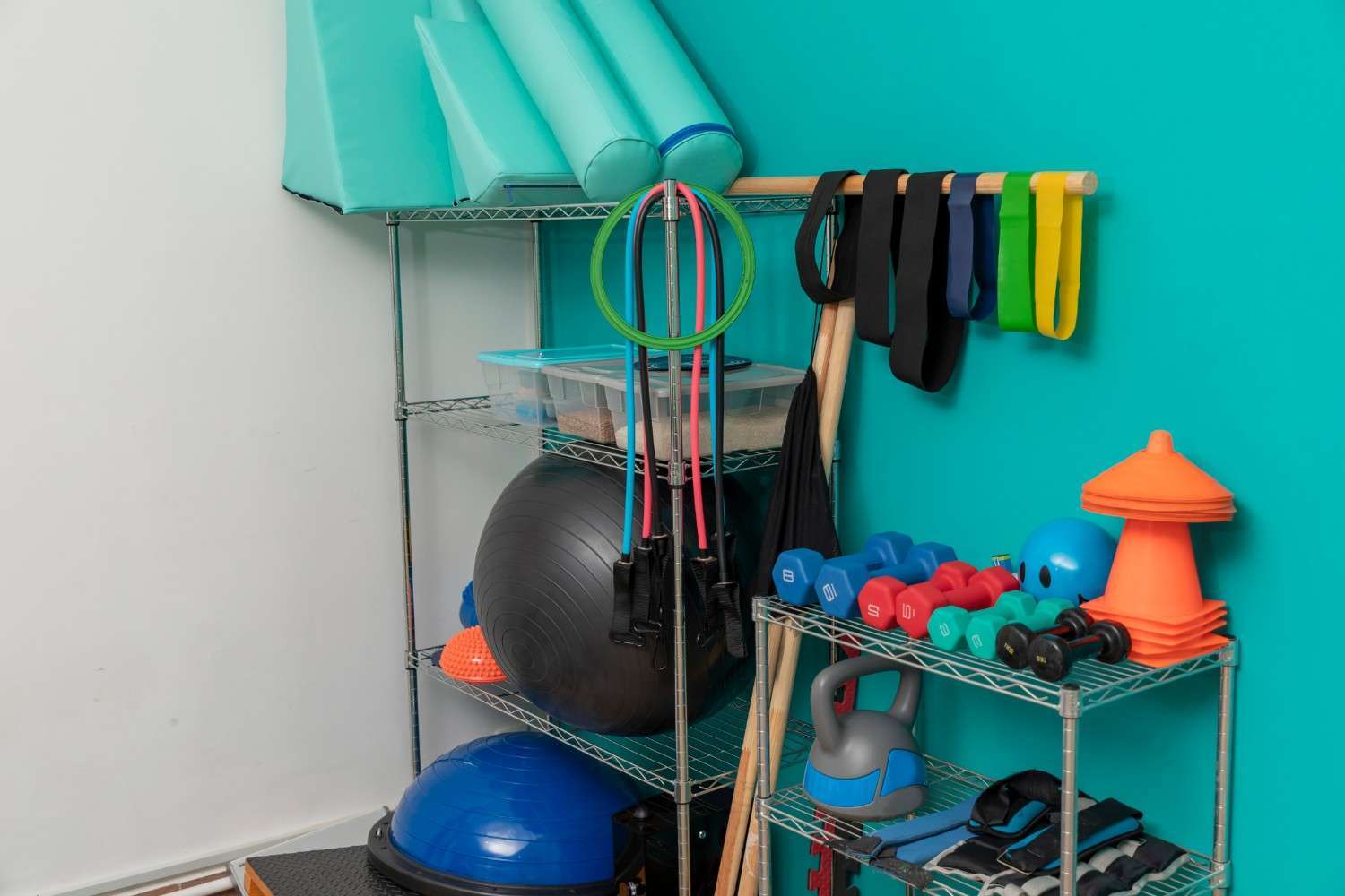 Photograph of a home gym corner featuring metal shelving units against a turquoise wall, holding various fitness equipment.