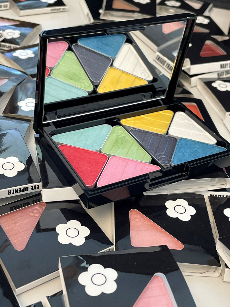 Eye Opener eyeshadows set into Colour Palette L. The palette is large and has a mirror and fits 8 triangular eye opener eye shadows inside. The palette is resting on single eyeshadows in their packaging- each is in a black cardboard sleeve with the Mary Quant Daisy on.