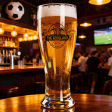 A beer glass filled with light golden beer, featuring a logo, set in a lively bar environment.