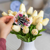 A hand holds a pink A7 greeting card featuring floral designs, placed among white tulips in a vase.