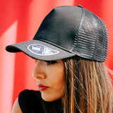 A black trucker cap with a mesh back and a logo on the front, worn by a model against a vibrant background.