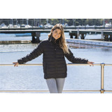 A woman wearing a black puffer jacket stands by the water, with a marina and bridge in the background.