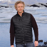 A man stands by a glacial lake wearing a black thermal vest over a long-sleeve top, with mountains in the background.