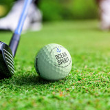 A green Pinnacle Soft golf ball rests on grass, with a golf club positioned nearby. The ball features a logo.