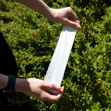 A person holds a white pet waste bag against a background of green foliage. The bag is flat and unused.