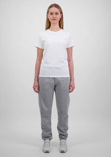 A woman wears a plain white t-shirt and grey track pants, standing against a light grey background. The pants have a logo.