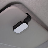 A car visor sunglass holder in black and white, mounted on a grey car interior. It has a simple, sleek design.