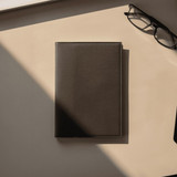 A dark brown leather refillable journal lies on a beige surface, accompanied by a pair of glasses.