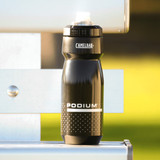A drink bottle in black with a grip design, featuring a logo and a nozzle cap on a metallic surface.