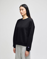 A black crewneck sweatshirt is displayed on a model, featuring a logo on the sleeve. The model wears grey pants.