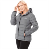 A woman wearing a grey puffer jacket with a hood and a logo on the front, standing with a hand on her shoulder.