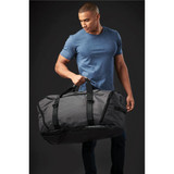A man holds a large grey duffle bag with black accents. The bag features short handles and a shoulder strap.