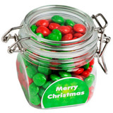 A glass canister filled with red and green chewy fruit candies, featuring a decorative label and a metal clasp.