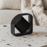 A black wireless speaker with a unique design sits on a speckled table, surrounded by a soft, textured background.