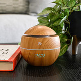 A wooden-patterned diffuser with a rounded design on a dark table, surrounded by greenery and a book.