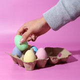 A hand reaching for a green pastel egg from a carton holding various pastel-coloured eggs on a pink background.