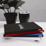 A set of three Pioneer RPET A5 Notebooks in black, red, and white, placed beside a plant and a drink.