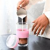 A reusable coffee cup in clear glass with a pink silicone sleeve, being filled with milk.