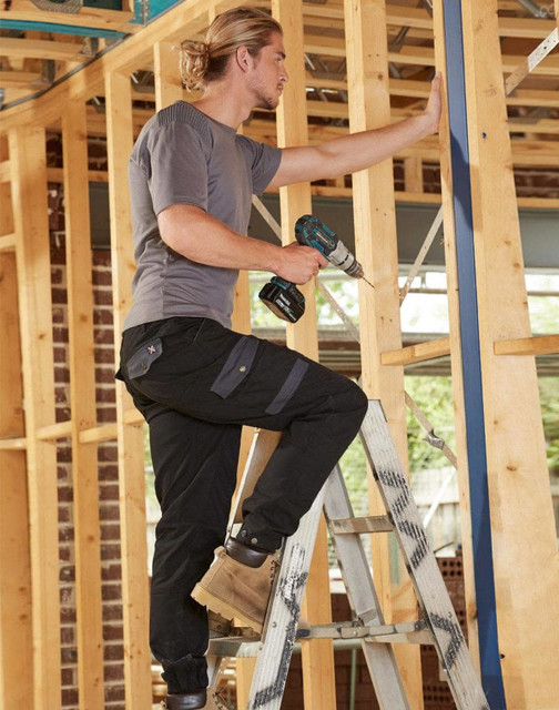 A pair of black work pants on a model using a power drill while standing on a ladder amidst wooden framing.