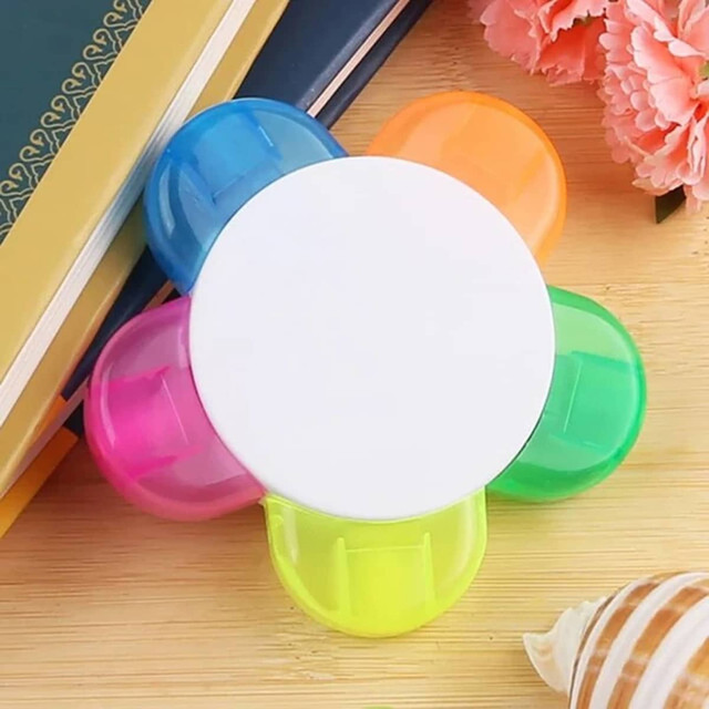 A flower-shaped highlighter with blue, orange, green, and yellow petals, sitting on a wooden surface with books and flowers.