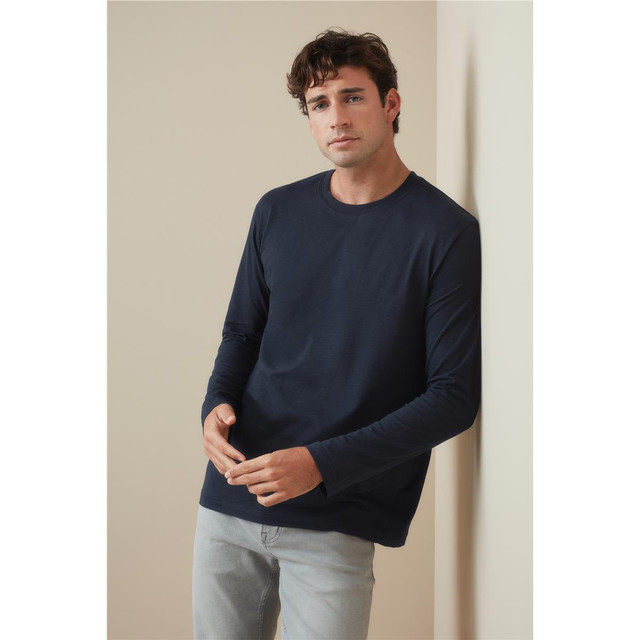 A man wearing a navy long-sleeve T-shirt leaning against a wall, paired with light grey trousers. The shirt features a logo.