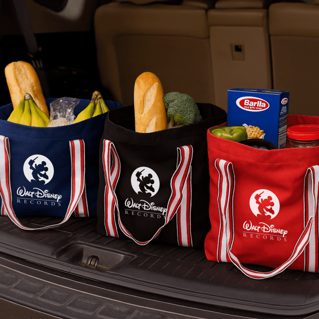 Three canvas tote bags in red, black, and blue, filled with groceries like bread, vegetables, and pasta. Each bag has a logo.