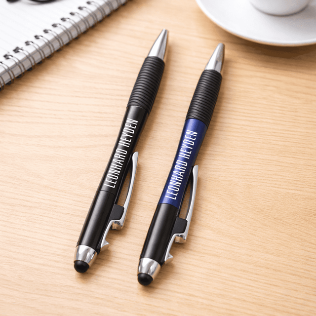 A pair of pens, one black and one blue, with grips, resting on a wooden surface near a cup and notebook.