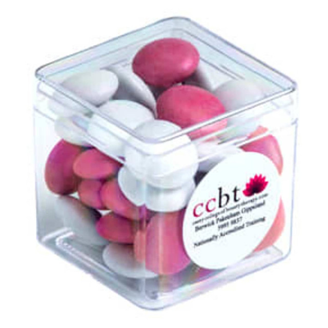 A clear cube container filled with pink and white chocolate-coated beans. The container has a label and branding.