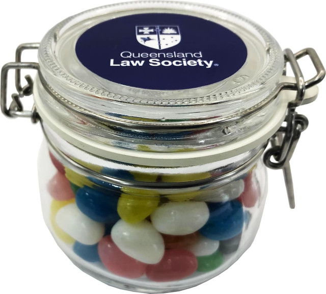 A clear glass canister filled with colourful mini jelly beans, featuring a decorative lid with a logo.