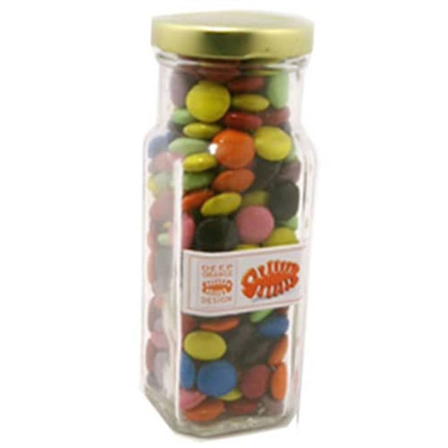 A glass tall jar filled with colourful choc beans, featuring a gold lid and a branding label.