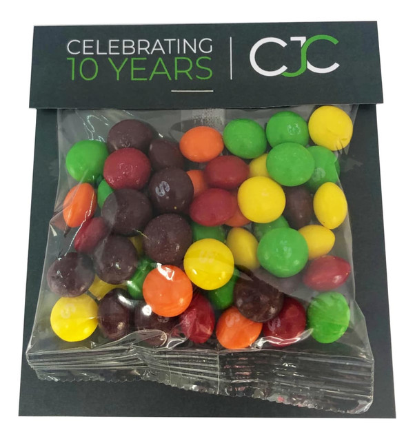 A transparent bag filled with colourful Skittles in red, yellow, green, orange, and purple, featuring a branded header.